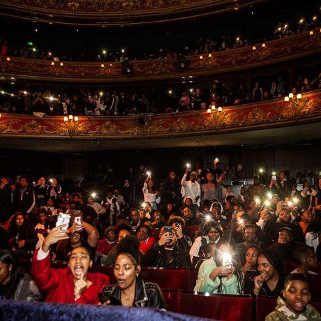 A photo of the Hackney Empire audience showing young people enjoying the act on stage with their camera lights on and held in the air.