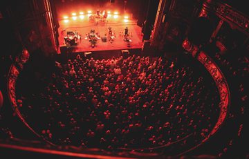 A photo taken from the higher levels of Hackney Empire showing Lankum playing on stage and a sold out audience watching them. There is a red light shining on the stage and onto the audience.