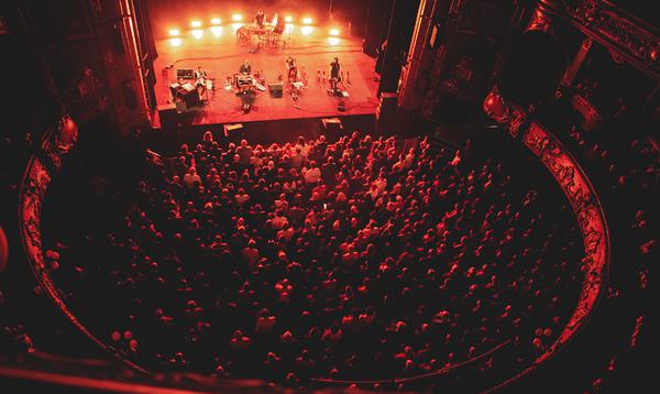 A photo taken from the higher levels of Hackney Empire showing Lankum playing on stage and a sold out audience watching them. There is a red light shining on the stage and onto the audience.