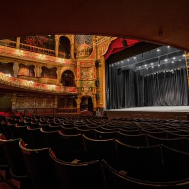 A photo taken from the back of the Stalls and underneath the overhang from the Dress Circle showing a view of the back of the seats and the blank stage.