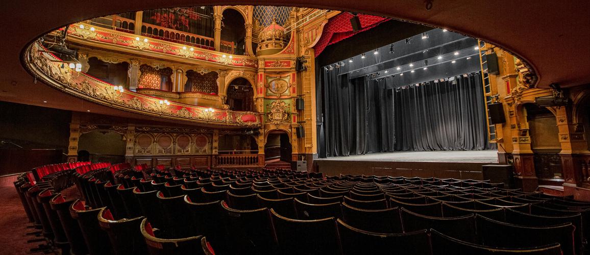 A photo taken from the back of the Stalls and underneath the overhang from the Dress Circle showing a view of the back of the seats and the blank stage.