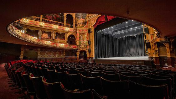 A photo taken from the back of the Stalls and underneath the overhang from the Dress Circle showing a view of the back of the seats and the blank stage.