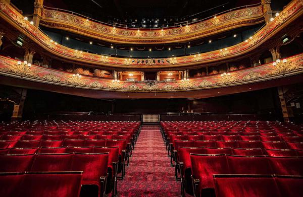 picture is taken from the front of the stalls showcasing the red seats in the stalls and the levels above with the house lights on.