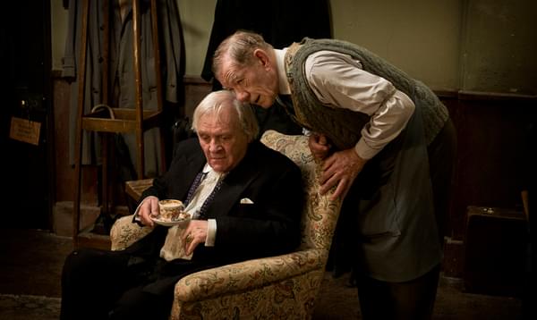 A photo of John Lithgow sat in a chair drinking tea whilst Ian McKellen leans over the back of the chair talking to him.