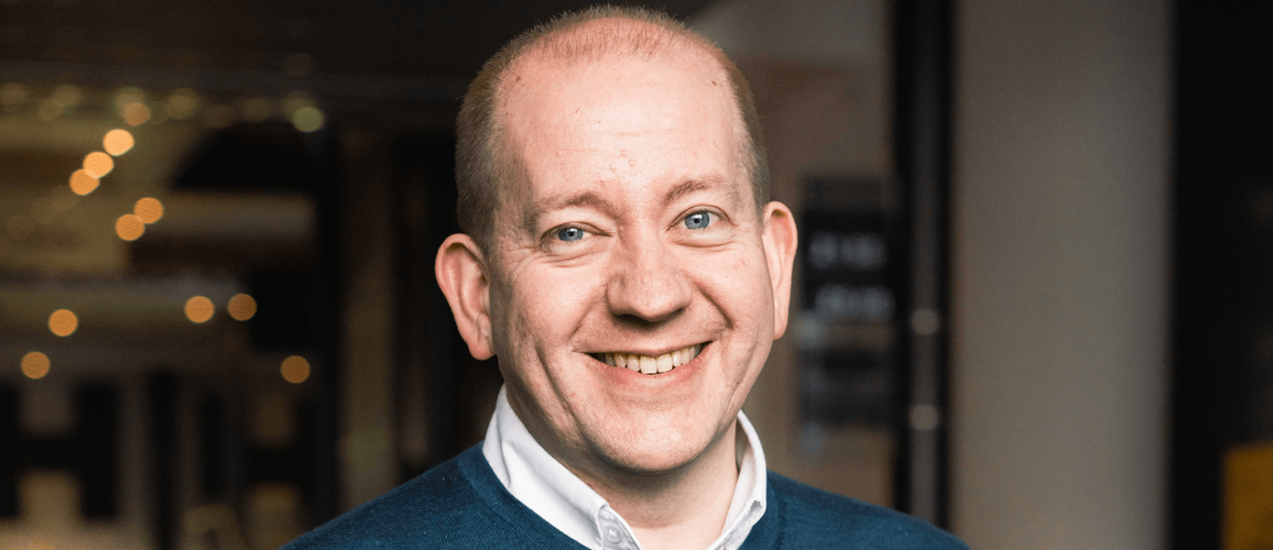 A photo of Chris Sudworth smiling at camera wearing a blue jumper over a shirt.