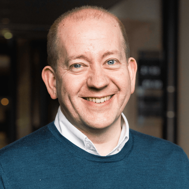 A photo of Chris Sudworth smiling at camera wearing a blue jumper over a shirt.