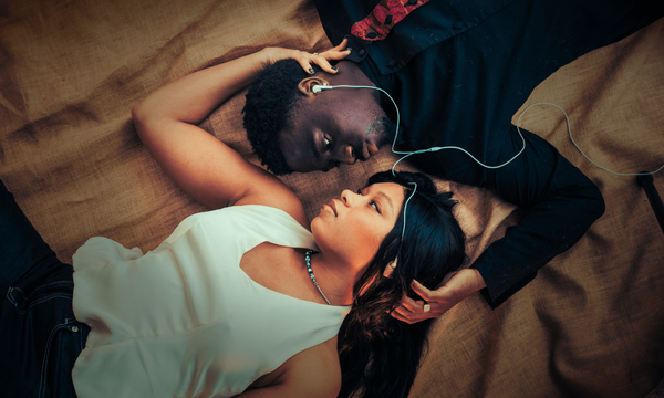 Two people lying on the ground with one arm round each others heads gazing at each other lovingly. They're wearing a headphone each which are connected.
