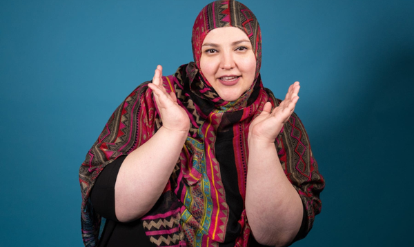 Fatiha El-Ghorri is standing against a blue background and hand her hands in the air cupping her face while smiling directly into the camera