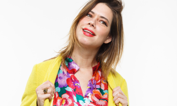Lou Sanders is wearing a colourful patterned dress with a yellow cardigan and is holding the sides of her cardigan while smiling at the camera