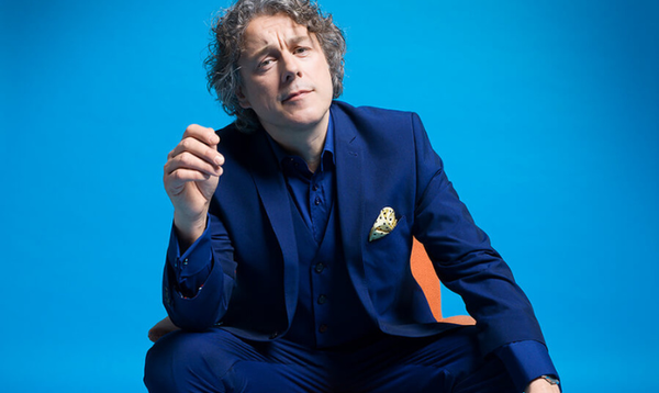 Alan Davies is wearing a blue suit, blue socks and bright yellow clogs on while sitting on a orange chair. He is looking straight at the camera with his right elbow resting on his knee