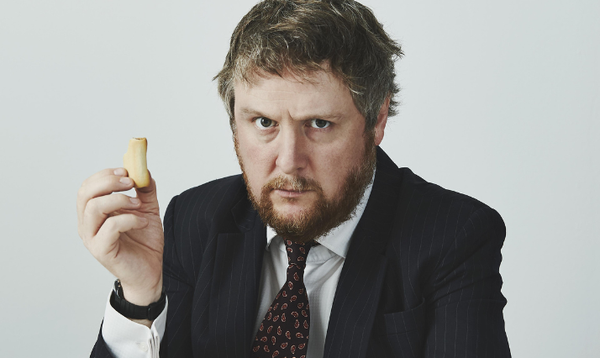 Tim Key is wearing a black suit and is holding a biscuit in his hands with an angry look on his face, while starting straight at the camera.