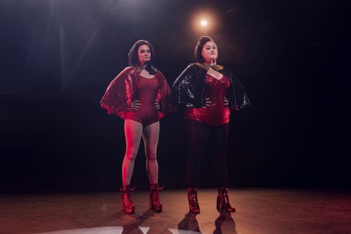 Rachel Fairburn and Kiri Pritchard-McLean stand side-by-side on a stage in glittery leotards, with heir hands on their hips, looking at the same spot in the distance. They are lit only by stage lights, which shine behind them.