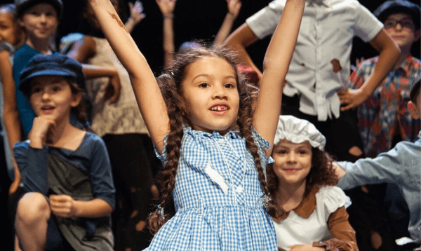 A young girl on stage in a spilt position with her hands in the air and is smiling at the audience