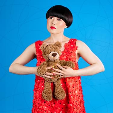 Atsuko Okatsuka is a Taiwan comedian with short jet black hair and is wearing a red top while holding a brown teddy bear.