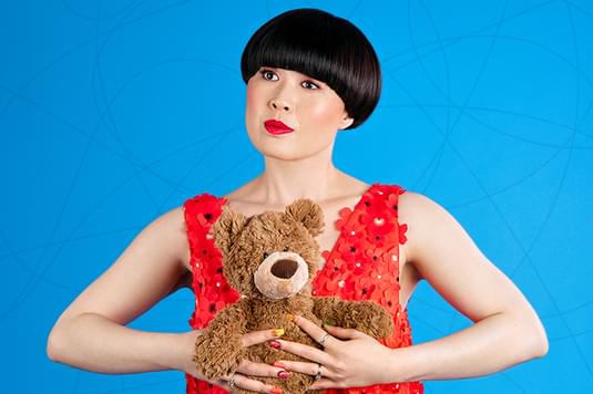 Atsuko Okatsuka is a Taiwan comedian with short jet black hair and is wearing a red top while holding a brown teddy bear.