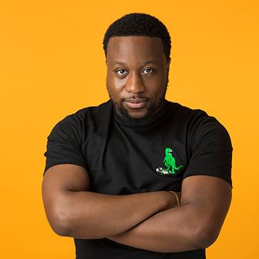 A photo of Babatunde Aléshé wearing a black t-shirt and staring at the camera, set against a yellow background.