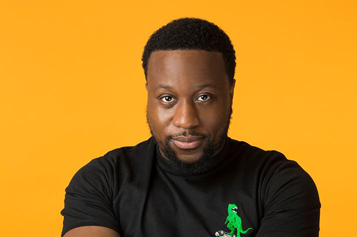 A photo of Babatunde Aléshé wearing a black t-shirt and staring at the camera, set against a yellow background.