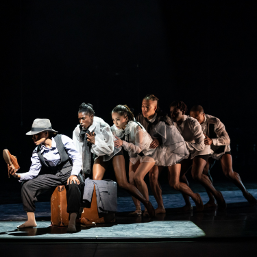 A group of dancers on stage creeping behind each other in a line while the one at the front of the line is sitting on a suitcase holding a shoe.