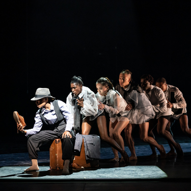 A group of dancers on stage creeping behind each other in a line while the one at the front of the line is sitting on a suitcase holding a shoe.