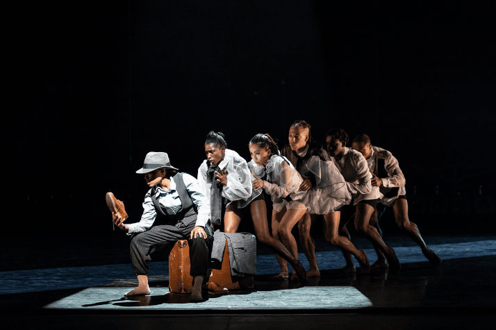 A group of dancers on stage creeping behind each other in a line while the one at the front of the line is sitting on a suitcase holding a shoe.
