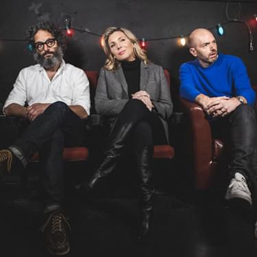 Paul Scheer, Jason Mantzoukas, and June Diane Raphael are sitting on a sofa all with their left eg crossed over their right leg and are all looking at the camera
