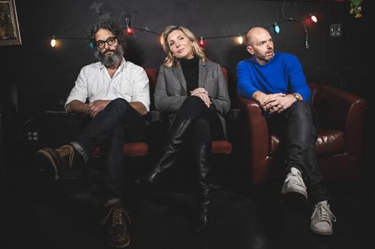 Paul Scheer, Jason Mantzoukas, and June Diane Raphael are sitting on a sofa all with their left eg crossed over their right leg and are all looking at the camera