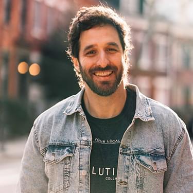 Rafi Bastos is a 6’7” tall Jewish-Brazilian comedian with dark brown hair and is wearing a black top and a Demin jacket while smiling straight at the camera.
