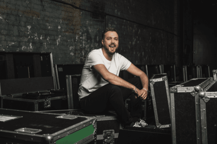 A photo of Iain Stirling smiling wearing a white t-shirt and sat on technical equipment