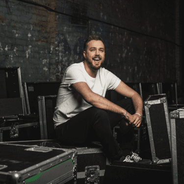 A photo of Iain Stirling smiling wearing a white t-shirt and sat on technical equipment
