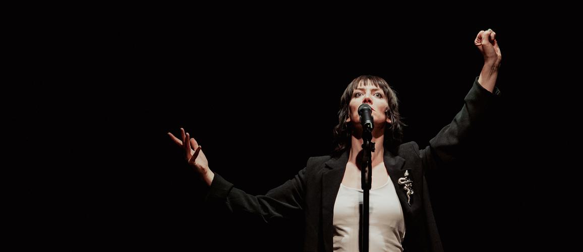 Actress Deborah Pugh is wearing a white vest and a black blazer with a snake brooch. She is standing in front of a mic stand with her hands up in the air. Photo taken by Camilla Adams.