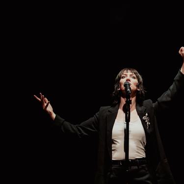 Actress Deborah Pugh is wearing a white vest and a black blazer with a snake brooch. She is standing in front of a mic stand with her hands up in the air. Photo taken by Camilla Adams.