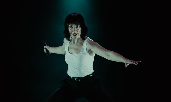 Actress Deborah Pugh is wearing a white vest and black trousers. She is facing directly at the camera and smiling. She has the mic in her right hand behind her body, and her left hand is towards the front. There is a pale green light on her