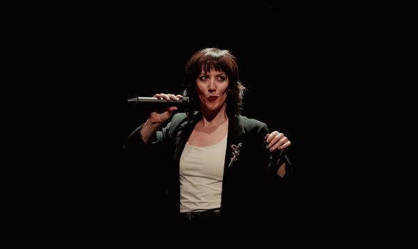 Actress Deborah Pugh is wearing a white vest and a black blazer with a snake brooch. She is holding the mic next to her lips with her right hand.