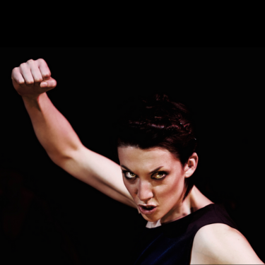 Actress Deborah Pugh is wearing a black vest and has her right fist punching up in the air. She is facing the camera with her head leaning forward. She is in front of a black background.