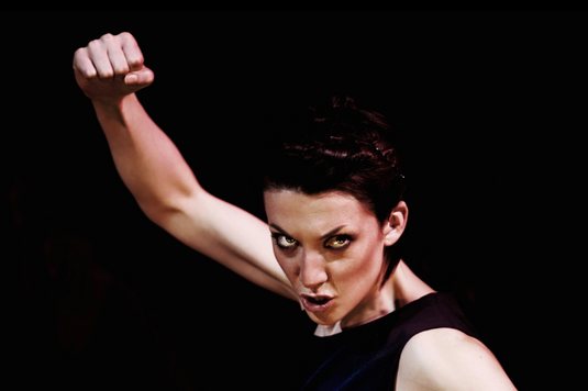 Actress Deborah Pugh is wearing a black vest and has her right fist punching up in the air. She is facing the camera with her head leaning forward. She is in front of a black background.