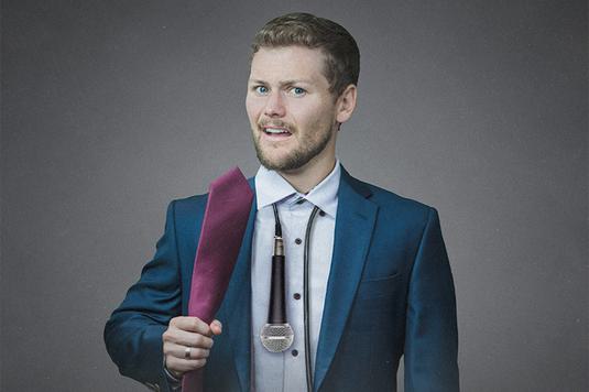 Drew Lynch wearing a blue suit with a microphone around his neck like a tie and holding a pink tie like a microphone.
