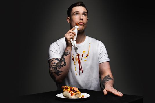 A photo of Ed Gamble in a white t-shirt with ketchup and mustard from a hot dog on the table all over his mouth and t-shirt.