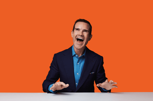 Jimmy Carr is wearing a navy blue suit with a baby blue shirt and is sitting at a table laughing with his hands in the air just above the table. there is also a orange background behind Jimmy Carr.
