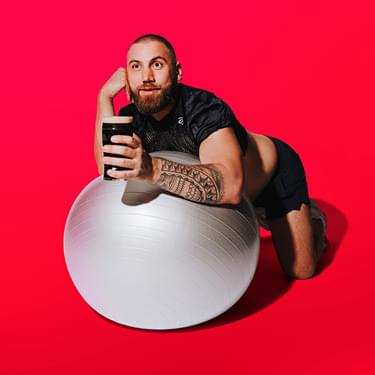 Morgan Rees leans forward on a silver exercise ball and looks into the distance. He's wearing a crop top and shorts. He rests his head on one hand and holds up a pint of Guiness up in the other. The background of the photo is a bright pink.