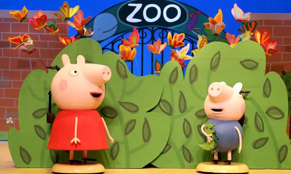 Peppa Pig and George Pig are standing outside of the zoo in front of the main entrance.