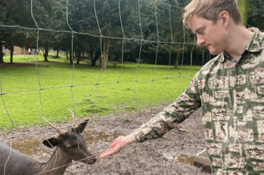 James Acaster is wearing a camouflage jacket and is feeding a goat on a farm