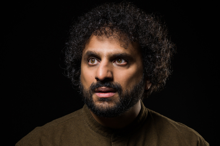 Nish Kumar is wearing a khaki shirt and is looking to his right away from the camera