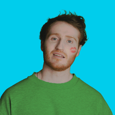 Ali Woods is wearing a green jumper and has a red lip kiss stain on his cheek and is standing against a bright blue background.