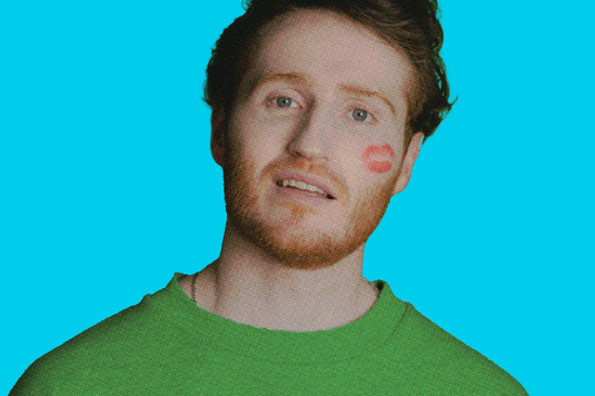 Ali Woods is wearing a green jumper and has a red lip kiss stain on his cheek and is standing against a bright blue background.