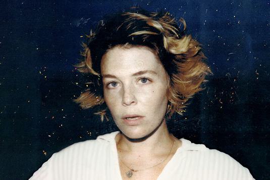 Maggie Rogers is staring at the camera with her mouth slightly open and a grainy effect on the photo, wearing a white blouse.