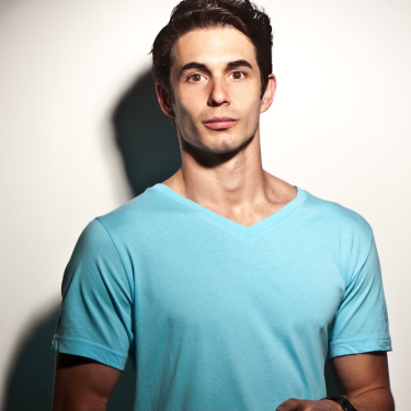 Michael Blaustein is wearing a bright blue v neck top and is standing against  white wall looking straight into the camera