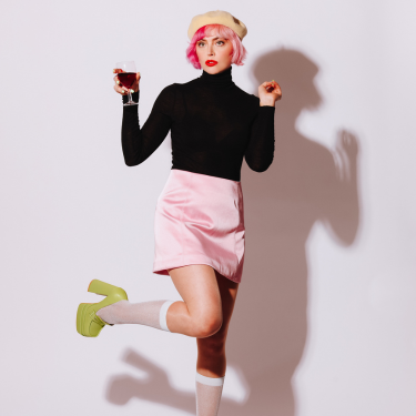 Tatty Mc Leod is wearing a bright pink beret hat with a black turtle neck and a pink skirt and is holding a glass in her hand.
