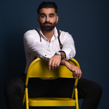 Tez is British Pakistani stand-up comedian, with short dark hair in a black tuxedo sitting on a yellow chair