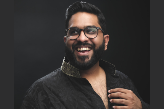 Eshaan Akbar, a man of South Asian heritage, is smiling and looking off camera. He is wearing a black shirt and glasses. He is in front of a black background.