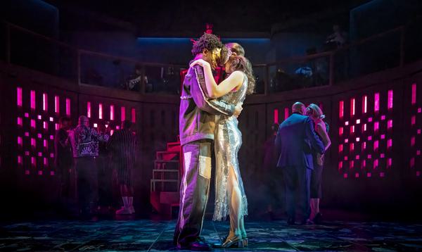 Kyle Ndukuba as Romeo and Mia Khan as Juliet stand arm in arm at the ball lit in a bright pink light.